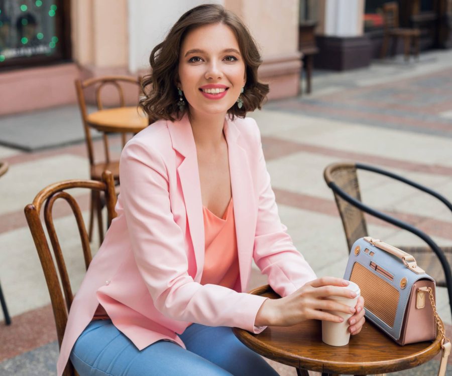 portrait of stylish smiling lady sitting at table drinking coffee in pink jacket summer style trend, blue handbag, accessories, street style, women fashion